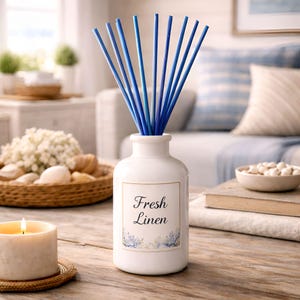 May include: A white ceramic essential oil diffuser with blue reeds. The label reads "Fresh Linen" with floral accents. A lit candle and a decorative bowl are in the foreground. The background includes a sofa with blue and white pillows.