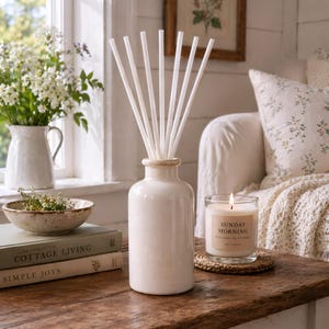 May include: A cream-colored ceramic diffuser with white reeds, a lit candle in a glass jar, and a small bowl with greenery sit on a wooden table. The candle label reads "Sunday Morning". A vase of flowers is in the background.
