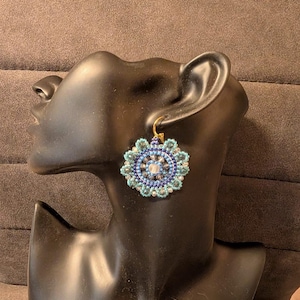 Può includere: Un orecchino circolare di perline con una perla centrale bianca circondata da anelli concentrici di perline blu scuro, blu chiaro e argento. Il bordo esterno è smerlato con perline blu chiaro e pende da un gancio in metallo dorato.