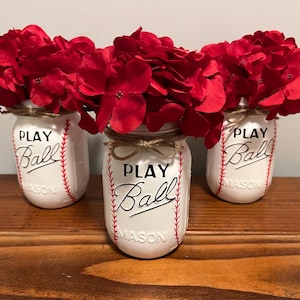 May include: Three white mason jars with red and white baseball stitching and the words "Play Ball" in black lettering. Each jar is filled with red hydrangea flowers.