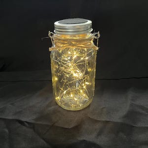 May include: A glass jar with a metal lid and a string of lights inside. The jar is decorated with twine and has a solar panel on the lid.