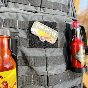 May include: A gray tactical backpack with two hot sauce bottles attached to the side. One bottle has a yellow label with a cartoon scorpion and the text "FUN METER" is on a patch on the front of the backpack.
