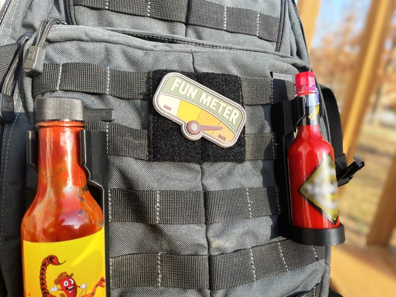 May include: A gray tactical backpack with two hot sauce bottles attached to the side. One bottle has a yellow label with a cartoon scorpion and the text "FUN METER" is on a patch on the front of the backpack.