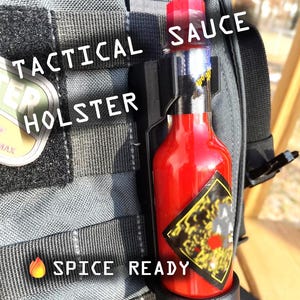 May include: A red hot sauce bottle with a black label and a black and gray tactical holster. The holster is attached to a gray tactical bag. The text "Tactical Holster" and "Spice Ready" are visible on the bag. The text "Sauce" is visible on the bottle.