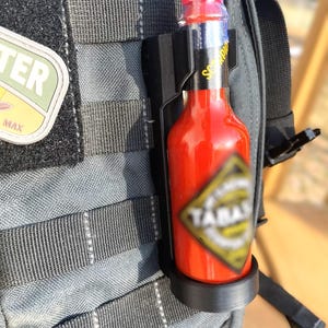 May include: A black hot sauce holder attached to a gray backpack. The holder is holding a red hot sauce bottle with a black and yellow label that says "Scorpion" and "Yamal".