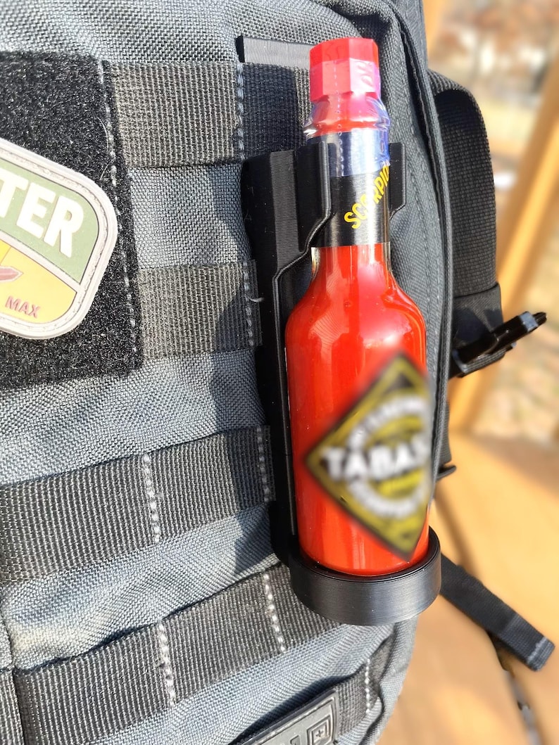 May include: A black hot sauce holder attached to a gray backpack. The holder is holding a red hot sauce bottle with a black and yellow label that says "Scorpion" and "Yamal".