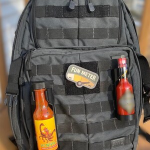 May include: A gray tactical backpack with two hot sauce bottles attached to the front. The bottles are in black holders and have labels with a scorpion and the text "POPPA'S". A patch with the text "FUN METER" is attached to the backpack.