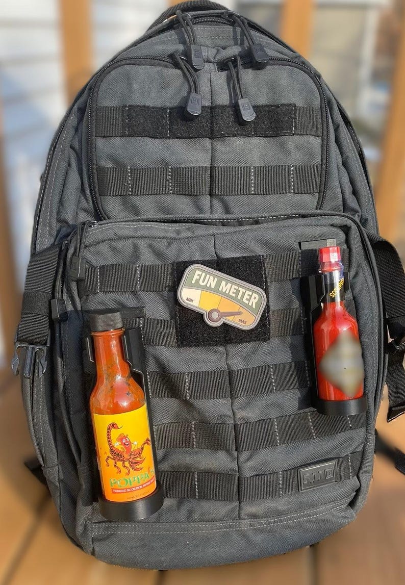 May include: A gray tactical backpack with two hot sauce bottles attached to the front. The bottles are in black holders and have labels with a scorpion and the text "POPPA'S". A patch with the text "FUN METER" is attached to the backpack.