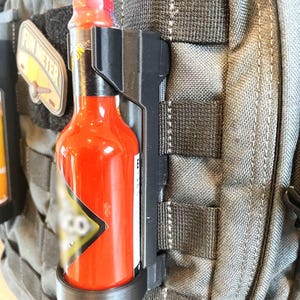 May include: A black plastic hot sauce holder attached to a gray backpack. The holder has a black strap and a black ring to hold the bottle. The hot sauce bottle is red and has a black label with white text.