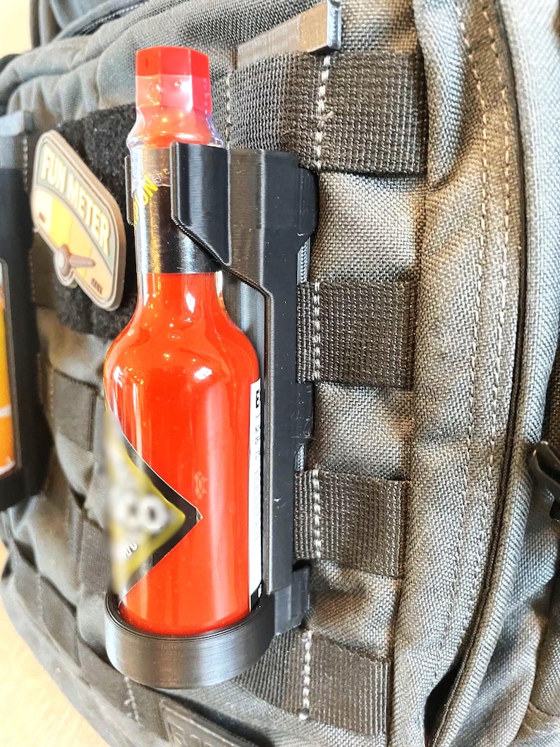 May include: A black plastic hot sauce holder attached to a gray backpack. The holder has a black strap and a black ring to hold the bottle. The hot sauce bottle is red and has a black label with white text.