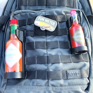 May include: A black tactical backpack with two red and black bottle holders. One bottle holder has a red hot sauce bottle with a white label. The other bottle holder has a red bottle with a black label. The backpack has a black patch with the text "FUN METER" on it.