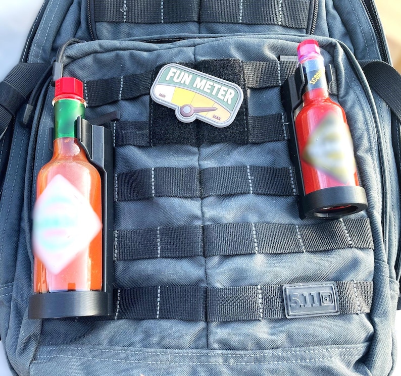 May include: A black tactical backpack with two red and black bottle holders. One bottle holder has a red hot sauce bottle with a white label. The other bottle holder has a red bottle with a black label. The backpack has a black patch with the text "FUN METER" on it.