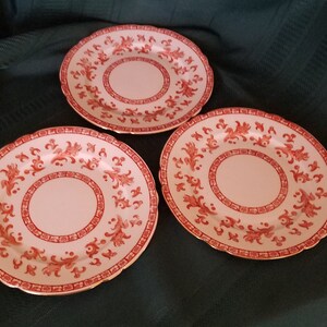May include: Three pink and white ceramic dinner plates with a red floral design and gold trim.