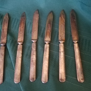 May include: Set of six silver butter knives with slightly rounded tips. The knives are laid out on a green fabric surface.