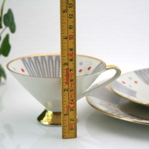 May include: A white teacup and saucer set with a geometric pattern in shades of grey and red. The cup has a gold rim and a gold base. The saucer has a gold rim. The set is 12.7 cm tall.
