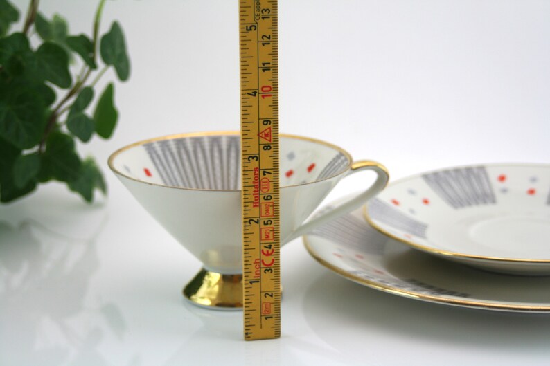 May include: A white teacup and saucer set with a geometric pattern in shades of grey and red. The cup has a gold rim and a gold base. The saucer has a gold rim. The set is 12.7 cm tall.