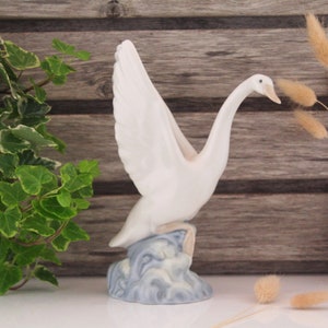 May include: White porcelain swan figurine with outstretched wings, standing on a blue and white base that resembles waves. The swan has a long neck and a small beak.