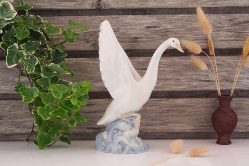 May include: White porcelain swan figurine with outstretched wings, standing on a blue and white base that resembles waves. The swan has a long neck and a small beak.