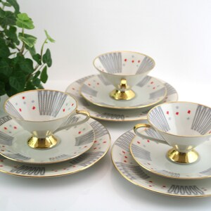 May include: Three vintage teacups and saucers featuring a geometric pattern in shades of grey and red. The cups have a gold rim and a gold base.