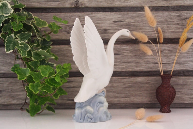 May include: White porcelain swan figurine with wings outstretched, standing on a blue and white base. The swan is perched on a wave-like base, suggesting it is in flight. The figurine is displayed on a wooden surface with a vase of dried grasses in the background.
