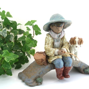 May include: A porcelain figurine of a young person wearing a blue hat and a brown jacket, sitting on a stone bridge with a dog. The figurine is holding a fishing rod and a basket.