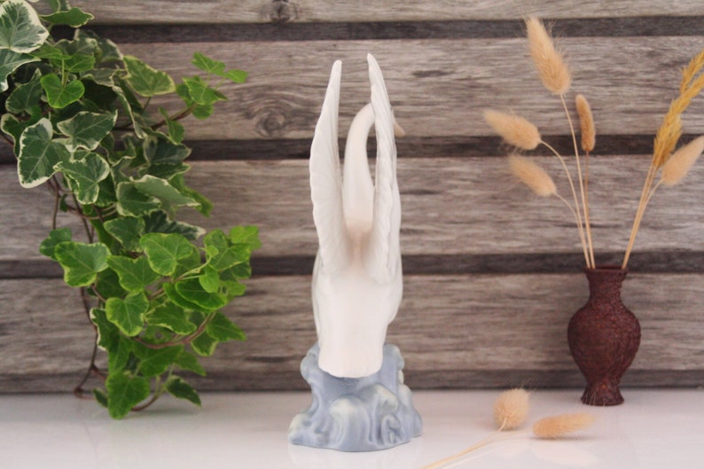 May include: White porcelain swan figurine with outstretched wings, standing on a blue and white base that resembles water waves.