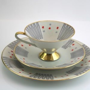 May include: A white teacup and saucer set with a geometric pattern in grey and red. The cup has a gold rim and a gold base. The saucer has a gold rim.