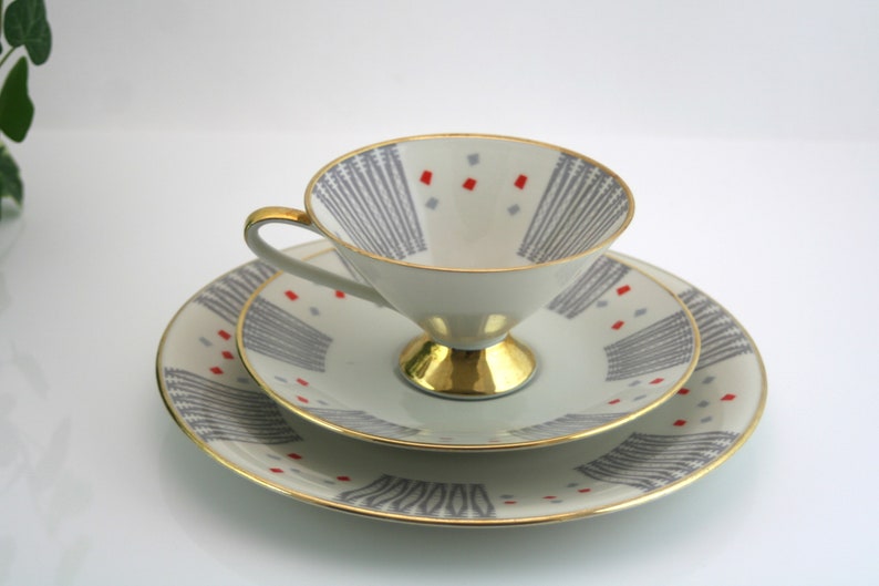 May include: A white teacup and saucer set with a geometric pattern in grey and red. The cup has a gold rim and a gold base. The saucer has a gold rim.