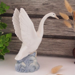 May include: A white porcelain swan figurine with outstretched wings, standing on a blue and white base that resembles water. The swan has a long neck and a small beak.
