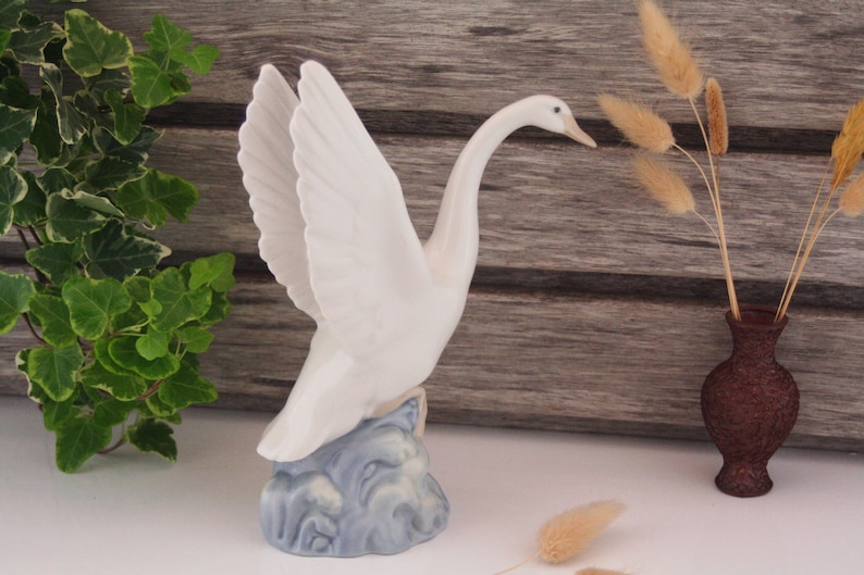 May include: A white porcelain swan figurine with outstretched wings, standing on a blue and white base that resembles water. The swan has a long neck and a small beak.