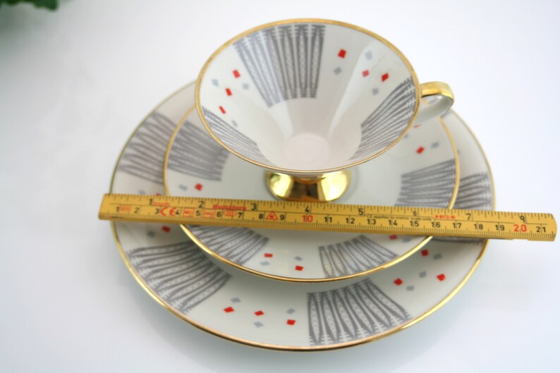May include: A set of three white porcelain teacups and saucers with a geometric pattern in shades of grey and red. The cups and saucers have a gold trim.