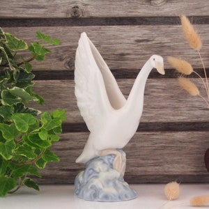 May include: White ceramic swan vase with outstretched wings, standing on a blue and white wave base. The vase is on a wooden surface with a green plant and dried grass in a brown vase.