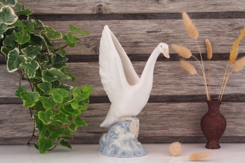 May include: White ceramic swan vase with outstretched wings, standing on a blue and white wave base. The vase is on a wooden surface with a green plant and dried grass in a brown vase.