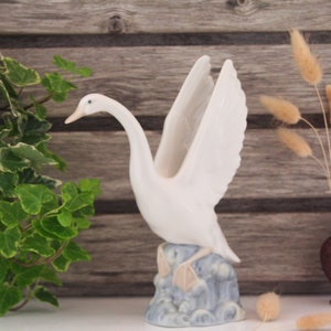 May include: White porcelain swan figurine with outstretched wings, standing on a blue and white base, with a leafy green plant and a brown vase with dried grasses in the background.