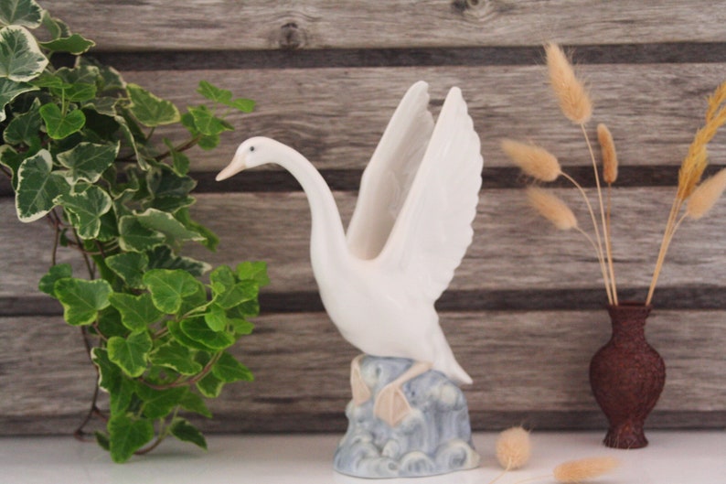 May include: White porcelain swan figurine with outstretched wings, standing on a blue and white base, with a leafy green plant and a brown vase with dried grasses in the background.