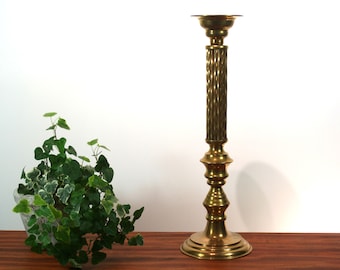 May include: A tall, gold-toned metal candlestick with a fluted column and a round base. The candlestick is on a wooden surface.