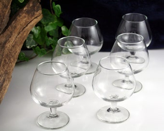 May include: Six clear glass brandy snifters with a round bowl and a short stem. The glasses are arranged in a semi-circle on a white surface.