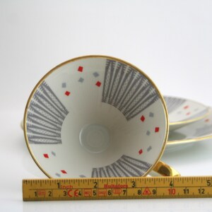 May include: A white teacup with a gold rim and a geometric pattern in grey and red. The cup is upside down and a ruler is in the foreground.