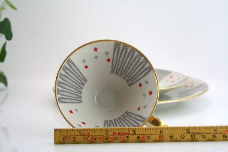 May include: A white teacup with a gold rim and a geometric pattern in grey and red. The cup is upside down and a ruler is in the foreground.