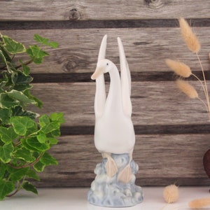 May include: White porcelain figurine of two swans with their necks intertwined, standing on a blue and white base. The swans are facing forward with their heads tilted up.