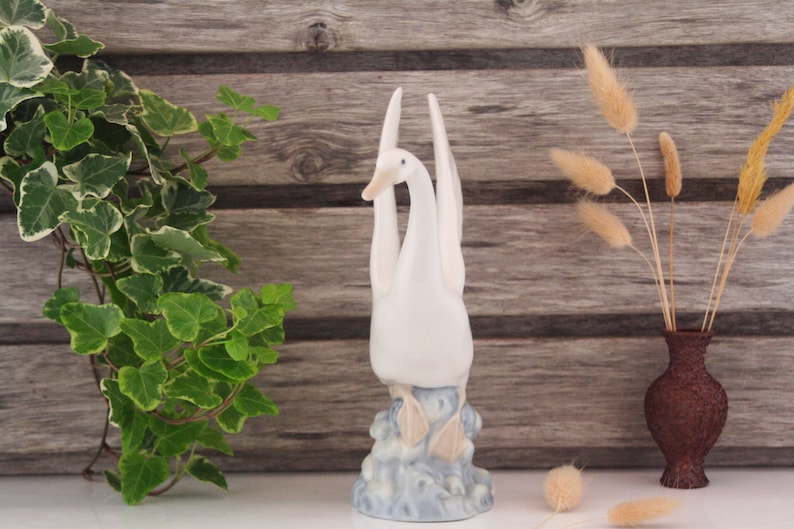 May include: White porcelain figurine of two swans with their necks intertwined, standing on a blue and white base. The swans are facing forward with their heads tilted up.