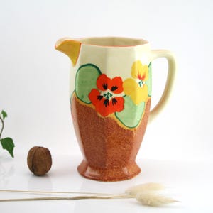 Op de afbeelding: Een vintage keramische kan met een geometrische vorm en een bloemenmotief. De kan is versierd met oranje, gele en groene bloemen op een crèmekleurige achtergrond. De kan heeft een bruine basis.