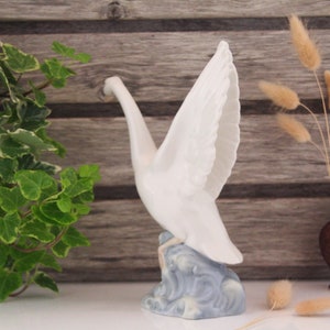 May include: White porcelain figurine of a crane with its wings spread, standing on a blue and white base. The crane is in a flying pose with its head tilted down.