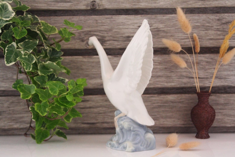 May include: White porcelain figurine of a crane with its wings spread, standing on a blue and white base. The crane is in a flying pose with its head tilted down.