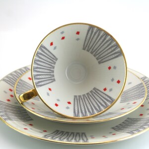 May include: A set of three white porcelain teacups and saucers with a geometric pattern in shades of grey and red. The cups and saucers have a gold rim.
