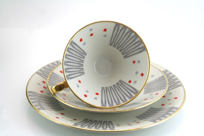 May include: A set of three white porcelain teacups and saucers with a geometric pattern in shades of grey and red. The cups and saucers have a gold rim.
