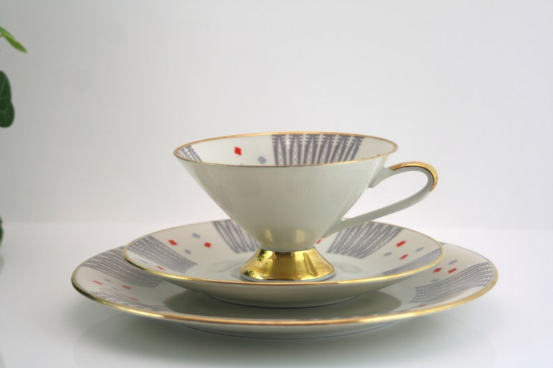May include: A white teacup and saucer set with a grey and red geometric pattern and gold trim.