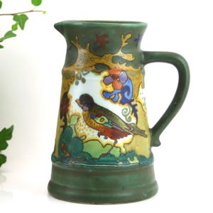Może przedstawiać: Zielony ceramiczny dzban z motywem ptaka i kwiatów. Dzbanek ma uchwyt i dzióbek. Ptak jest przedstawiony z czerwonymi, pomarańczowymi i zielonymi piórami. Dzbanek ma około 20 cm wysokości.
