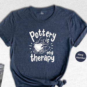 May include: A navy blue t-shirt with the text "Pottery is my therapy" in white. The text is accompanied by a graphic of a hand shaping a clay pot on a pottery wheel.