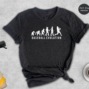 May include: Dark grey heather t-shirt with a white graphic of a baseball evolution. The graphic shows a series of stick figures evolving from a caveman to a baseball player. The text "BASEBALL EVOLUTION" is printed below the graphic.